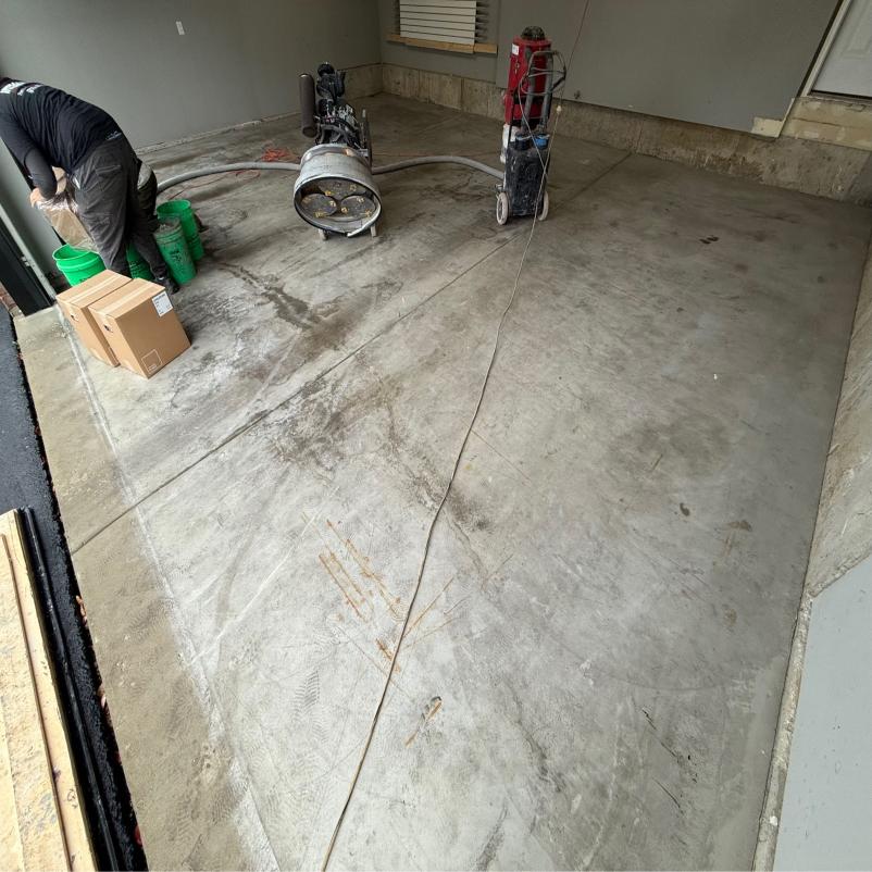 A person is working with concrete-grinding equipment in a garage, alongside buckets and boxes. The floor is partially polished.