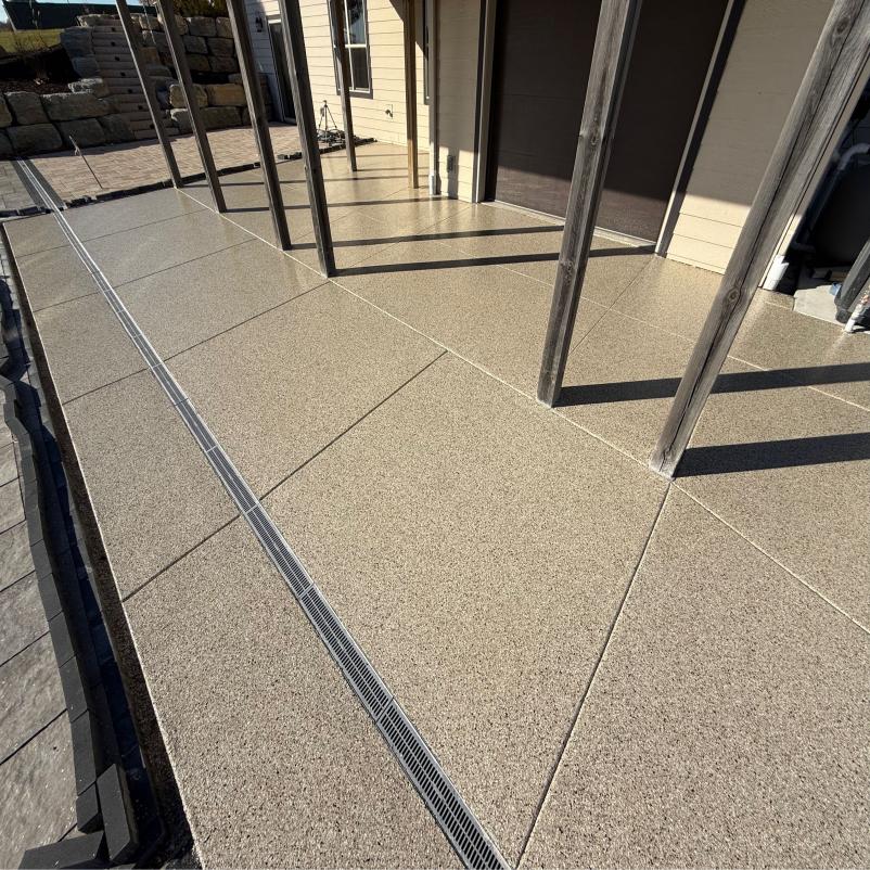 Modern patio with large, polished beige tiles and wooden posts. The area is clean and sunlit, with a drainage strip along the tiles.