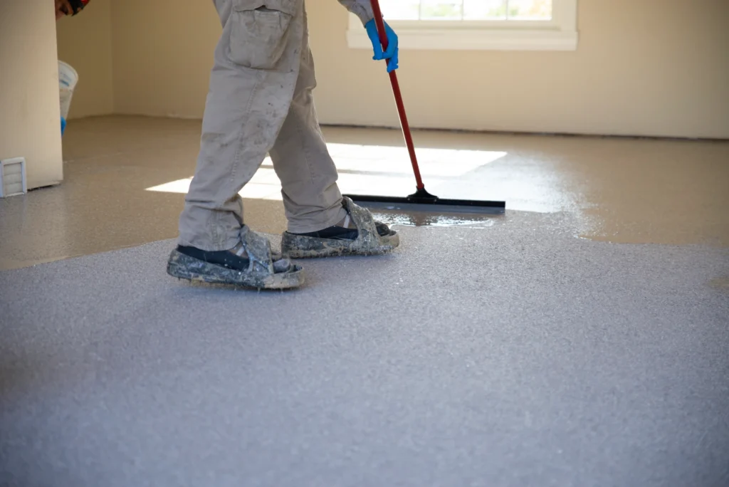 A person using a squeegee on a wet, gray floor, wearing protective clothing and overshoes, with sunlight entering through a window.